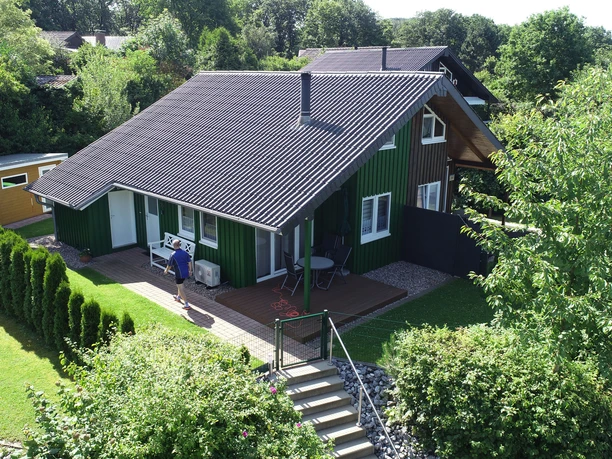 Grünes Holzhaus mit Terrasse, umgeben von Garten und Bäumen, Menschen genießen die Sommeratmosphäre.