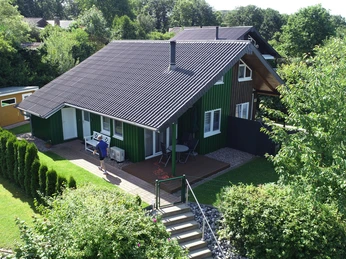 Ferienpark Extertal_Haus Luna Grünes Holzhaus mit Terrasse, umgeben von Garten und Bäumen, Menschen genießen die Sommeratmosphäre.