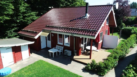 Ferienpark Extertal_Haus Mara Ferienhaus aus Holz mit rotem Dach und Terrasse, umgeben von grüner Rasenfläche und Bäumen.