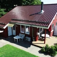 Ferienpark Extertal_Haus Mara Ferienhaus aus Holz mit rotem Dach und Terrasse, umgeben von grüner Rasenfläche und Bäumen.