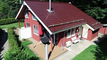 Ferienpark Extertal_Haus Mia Rot gestrichenes Holzhaus mit rotem Satteldach, große Terrasse, Grill und umgeben von Bäumen.