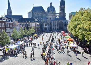 Blick auf den Aachener Dom und den Katschhof, auf dem zwei Stände und viele Tische, an denen Personen sitzen oder stehen, aneinander gereiht sind.