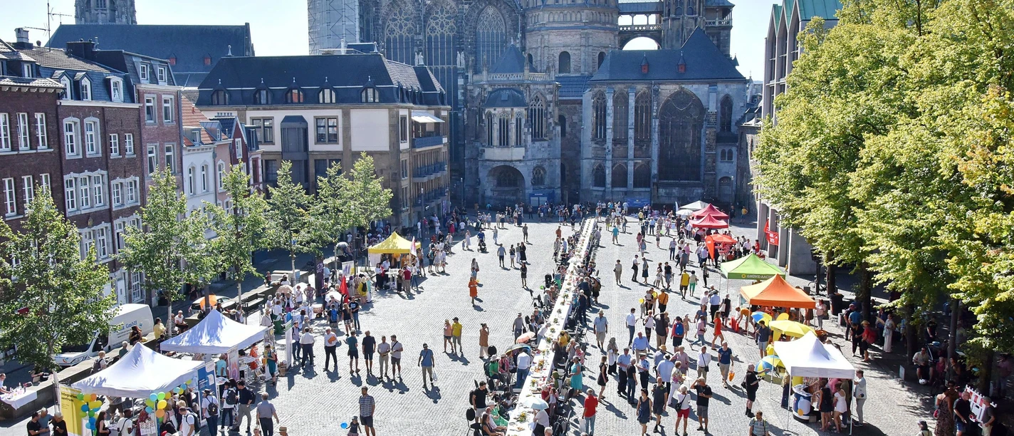 Lange Tafel der Vielfalt_51.jpg Blick auf den Aachener Dom und den Katschhof, auf dem zwei Stände und viele Tische, an denen Personen sitzen oder stehen, aneinander gereiht sind.