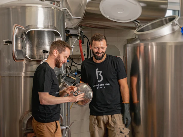 Zwei Männer stehen in einer Brauerei zwischen großen silbernen Behältern und lachen.