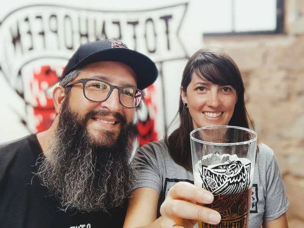 Eine Frau hält ein gefülltes Bierglas in der Hand und neben ihr steht ein Mann mit langem Bart, Brille und Kappe. Beide lächeln in die Kamera.