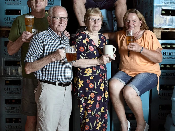 Zwei Frauen und drei Männer unterschiedlichen Alters stehen zwischen hoch gestapelten Bierkästen und halten entweder ein Bierglas, -krug oder -flaschen in der Hand.