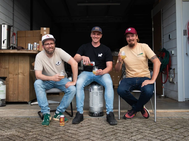 AllezHop6.jpg Drei Männer sitzen nebeneinander, lächeln in die Kamera und halten Biergläser in der Hand.