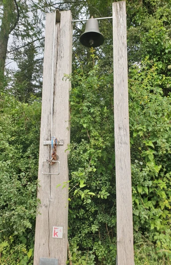 Zwei hohe Holzbalken mit Glocke in waldreicher Umgebung, umgeben von dichtem Grün und Bäumen.