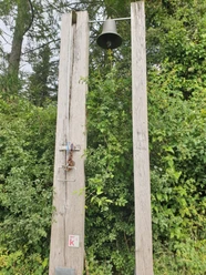Klima-Erlebnis-Route Zwei hohe Holzbalken mit Glocke in waldreicher Umgebung, umgeben von dichtem Grün und Bäumen.