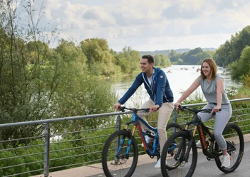 Werre-Weser-Kuss Zwei Personen radeln fröhlich auf einem Uferweg entlang eines Flusses, von Natur umgeben.