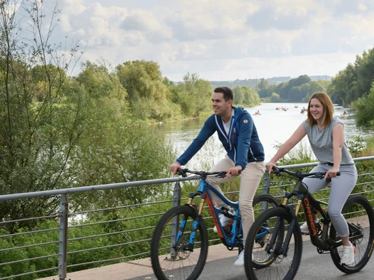 Werre-Weser-Kuss Zwei Personen radeln fröhlich auf einem Uferweg entlang eines Flusses, von Natur umgeben.