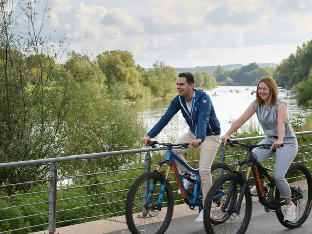 Werre-Weser-Kuss Zwei Personen radeln fröhlich auf einem Uferweg entlang eines Flusses, von Natur umgeben.