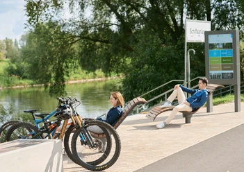 Werre-Weser-Kuss Ruhebänke Zwei Menschen entspannen auf modernen Ruhebänken nahe eines Flusses, mit geparkten Fahrrädern in der Nähe.