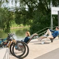 Werre-Weser-Kuss Ruhebänke Zwei Menschen entspannen auf modernen Ruhebänken nahe eines Flusses, mit geparkten Fahrrädern in der Nähe.