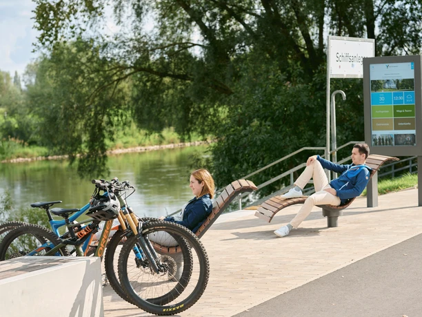 Werre-Weser-Kuss Ruhebänke Zwei Menschen entspannen auf modernen Ruhebänken nahe eines Flusses, mit geparkten Fahrrädern in der Nähe.