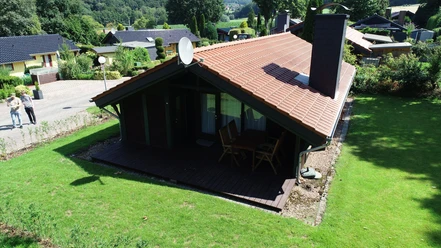 Ferienpark Extertal_Haus Wiesel Ferienhaus im Grünen mit rotem Dach und Veranda, umgeben von Rasen und weiteren Ferienhäusern.