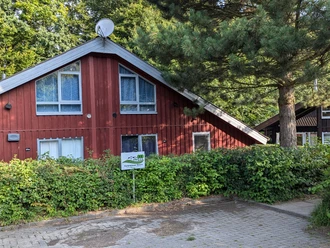 Ferienpark Extertal_Komfort Ferienhaus Rot Rotes Ferienhaus mit Sat-Schüssel, umgeben von Bäumen und Sträuchern, Parkplatz vorne.