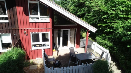 Ferienpark Extertal_Komfort-Ferienhaus Sonnental Rotes Holzferienhaus mit Terrasse und Gartenmöbeln, umgeben von hoher, grüner Vegetation.