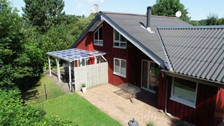Ferienpark Extertal_Sonnenhaus Rotes Ferienhaus mit grauem Dach, umgeben von Bäumen, Terrasse mit Glasüberdachung, sonnig.