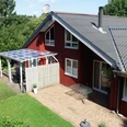 Ferienpark Extertal_Sonnenhaus Rotes Ferienhaus mit grauem Dach, umgeben von Bäumen, Terrasse mit Glasüberdachung, sonnig.