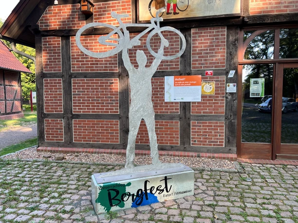 A sculpture of a cyclist holding up a bicycle, in front of a half-timbered building on cobblestones.