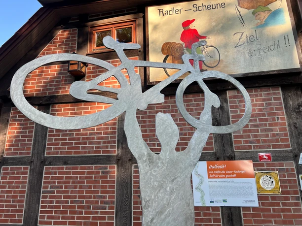 A metal sculpture of a person holding a bicycle above their head, in front of a brick-clad wall.