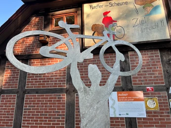 Skulptur Bergfest Metallskulptur einer Person, die ein Fahrrad über dem Kopf hält, vor einer mit Backstein verkleideten Wand.