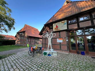 Skulptur eines Radfahrers mit erhobenem Fahrrad vor einem Fachwerkgebäude mit Fahrradständer.