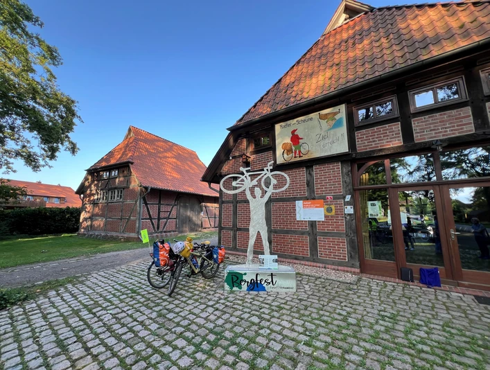 Skulptur Bergfest Skulptur eines Radfahrers mit erhobenem Fahrrad vor einem Fachwerkgebäude mit Fahrradständer.