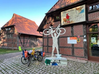 Metallskulptur eines Radfahrers mit Fahrrad über Kopf vor Fachwerkgebäude und Radscheune-Schild.