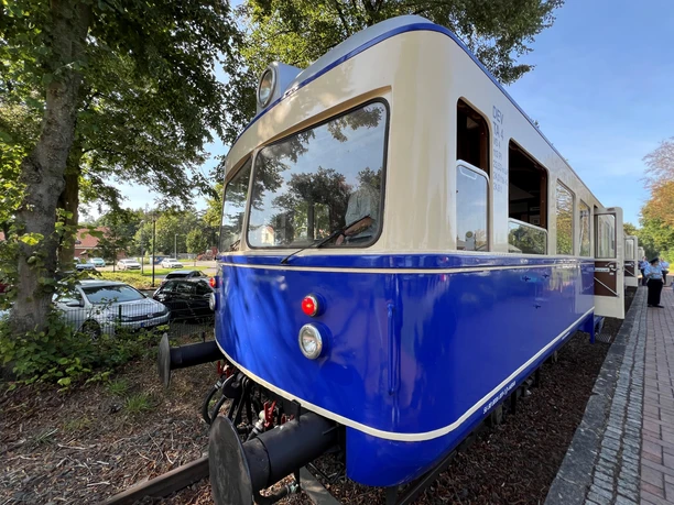 A blue vintage railbus is parked on a platform, surrounded by trees and parking spaces.
