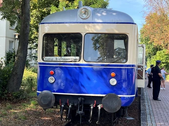 Ein historischer, blauer Nahverkehrszug, Kaffkieker, steht auf einem Gleis neben Spaziergängern im Grünen.