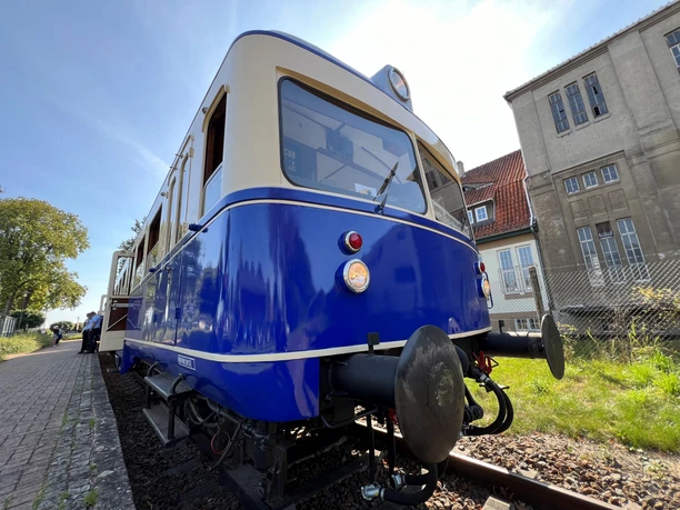 Ein blau-weißer Zug steht auf Gleisen, umgeben von historischen Gebäuden und einer Grünfläche.