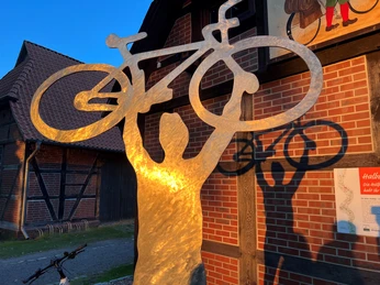 Skulptur aus Metall mit einem Radfahrer, gehalten von Armen über Kopf, reflektiert im Abendlicht.