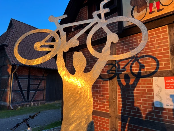 Skulptur aus Metall mit einem Radfahrer, gehalten von Armen über Kopf, reflektiert im Abendlicht.