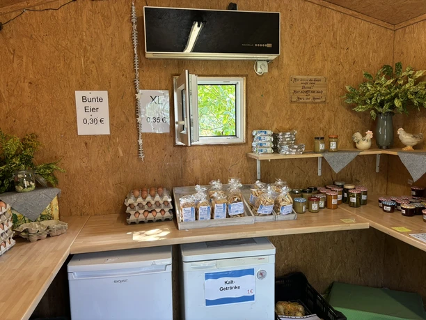 Innenraum eines kleinen Hofladens mit Eiern, Marmelade, Gebäck, Pflanzen und Kühlschränken.