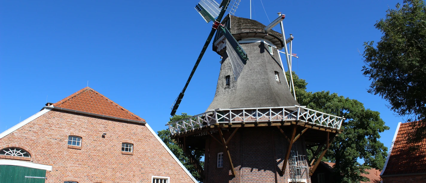 Mühle Hahnentange, Rhauderfehn Mühle Hahnentange mit Reetdach und Galerie neben rotem Backsteinhaus vor blauem Himmel.