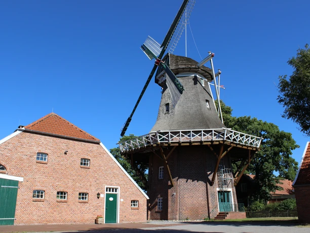 Mühle Hahnentange, Rhauderfehn Mühle Hahnentange mit Reetdach und Galerie neben rotem Backsteinhaus vor blauem Himmel.