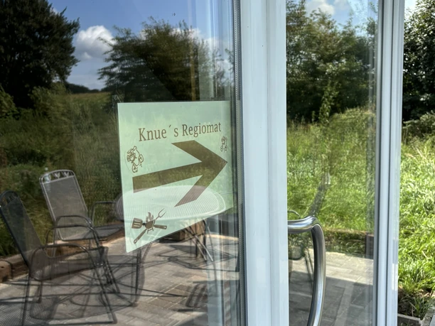 Knues Regiomat ©Emsland Tourismus GmbH Hinweisschild zu Knue’s Regiomat an einer Glastür mit Spiegelung von Gartenmöbeln und grüner Wiese