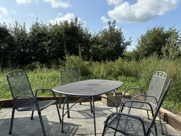 Knues Regiomat ©Emsland Tourismus GmbH Metalltisch mit vier Stühlen auf Terrasse vor grüner Wiese und Bäumen unter blauem Himmel.