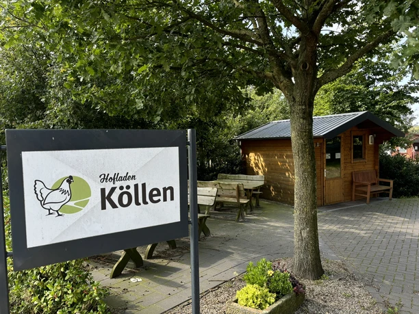Hofladen Köllen Holzhaus mit Sitzbänken und Schild des Hofladens Köllen unter einem großen belaubten Baum