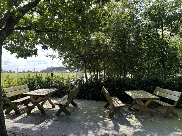 Hofladen Köllen Holzbänke und Tische im Schatten grüner Bäume mit Blick auf eine weite Wiesenlandschaft.