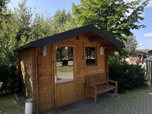 Hofladen Köllen Kleines Holzhaus mit Bank davor, umgeben von Bäumen und Bauernhofgebäuden bei sonnigem Wetter.