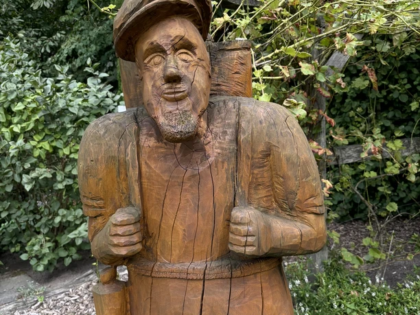 Bibelgarten Salzbergen ©Emsland Tourismus GmbH Holzfigur eines bärtigen Mannes mit Rucksack und Mütze, umgeben von grünem Laub im Bibelgarten.