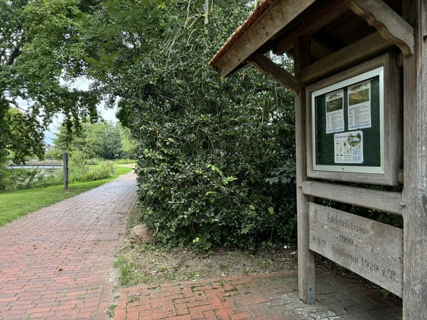 Fischereilehrpfad ©Emsland Tourismus GmbH Holztafel mit Infomaterial am Fischereilehrpfad neben gepflastertem Weg und grüner Uferbepflanzung.