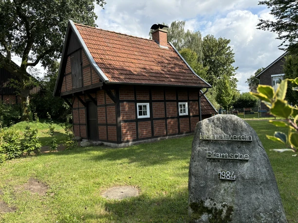 Heimathof Bramsche ©Emsland Tourismus GmbH Kleines Fachwerkhaus mit rotem Ziegeldach auf grüner Wiese, davor ein Gedenkstein mit Inschrift.