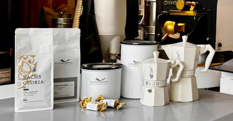 Produits du torréfacteur de café Elstermühle - Produits régionaux de Leipzig Les différents produits du torréfacteur de café Elstermühle de Leipzig, dont des grains de café frais emballés, des récipients de conservation et des cafetières espresso, sont exposés devant les machines à torréfier le café.