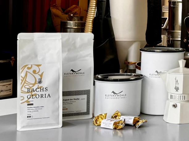Produkte der Elstermühle Kaffeerösterei - Regionale Produkte aus Leipzig Auf einer Auslage vor Kaffeeröstmaschinen stehen die verschiedenen Produkte der Elstermühle Kaffeerösterei aus Leipzig, darunter frische verpackte Kaffeebohnen, Aufbewahrungsbehälter und Espressokocher
