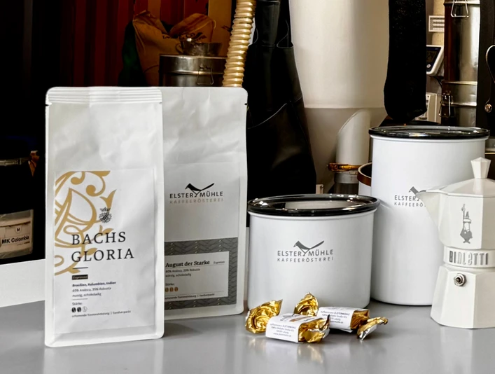 Produkte der Elstermühle Kaffeerösterei - Regionale Produkte aus Leipzig Auf einer Auslage vor Kaffeeröstmaschinen stehen die verschiedenen Produkte der Elstermühle Kaffeerösterei aus Leipzig, darunter frische verpackte Kaffeebohnen, Aufbewahrungsbehälter und Espressokocher