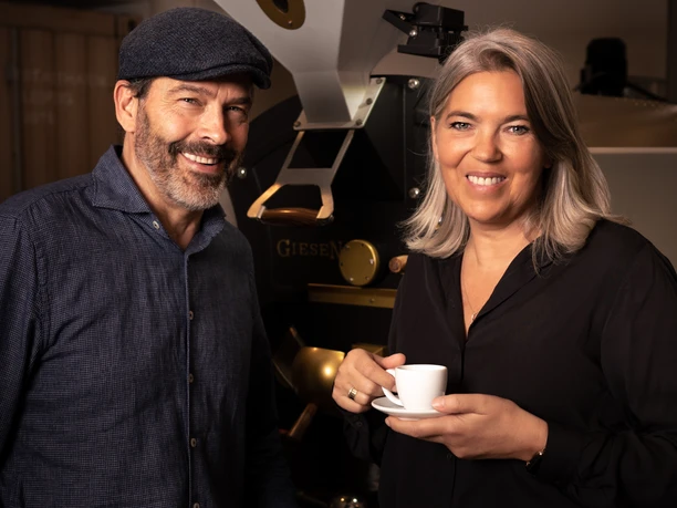Mario and Annette Rose from the Elstermühle coffee roastery - regional products from Leipzig Portrait of the owners of the Elstermühle coffee roastery, Mario and Annette Rose, in front of a coffee roasting machine in their roastery. Annette Rose holds a coffee cup in her hand